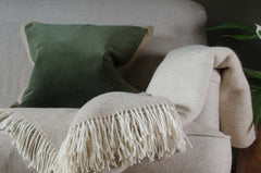 Beige sofa with a green velvet cushion, next to a reclaimed wooden coffee table and potted plant and beige pure wool blanket
