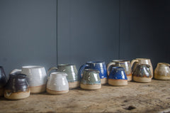Row of ceramic handmade mini milk jugs in green & mugs in various colours on a wooden surface with a dark grey background