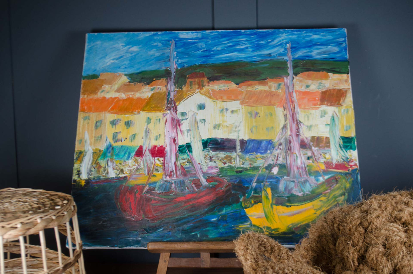 A colorful oil painting of a coastal harbor with sailboats, displayed on a wooden easel against a dark gray wall. The painting features vibrant blues, reds, yellows, and oranges, with a woven wicker basket and a textured brown rug partially visible in the foreground.