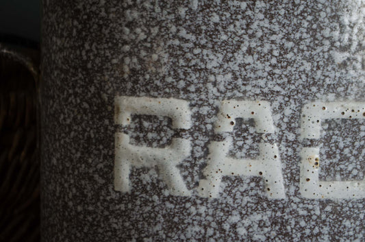 Close-up of a speckled ceramic mug or container with a brown and white marbled glaze and the letters 'RAC' in white raised on the front.