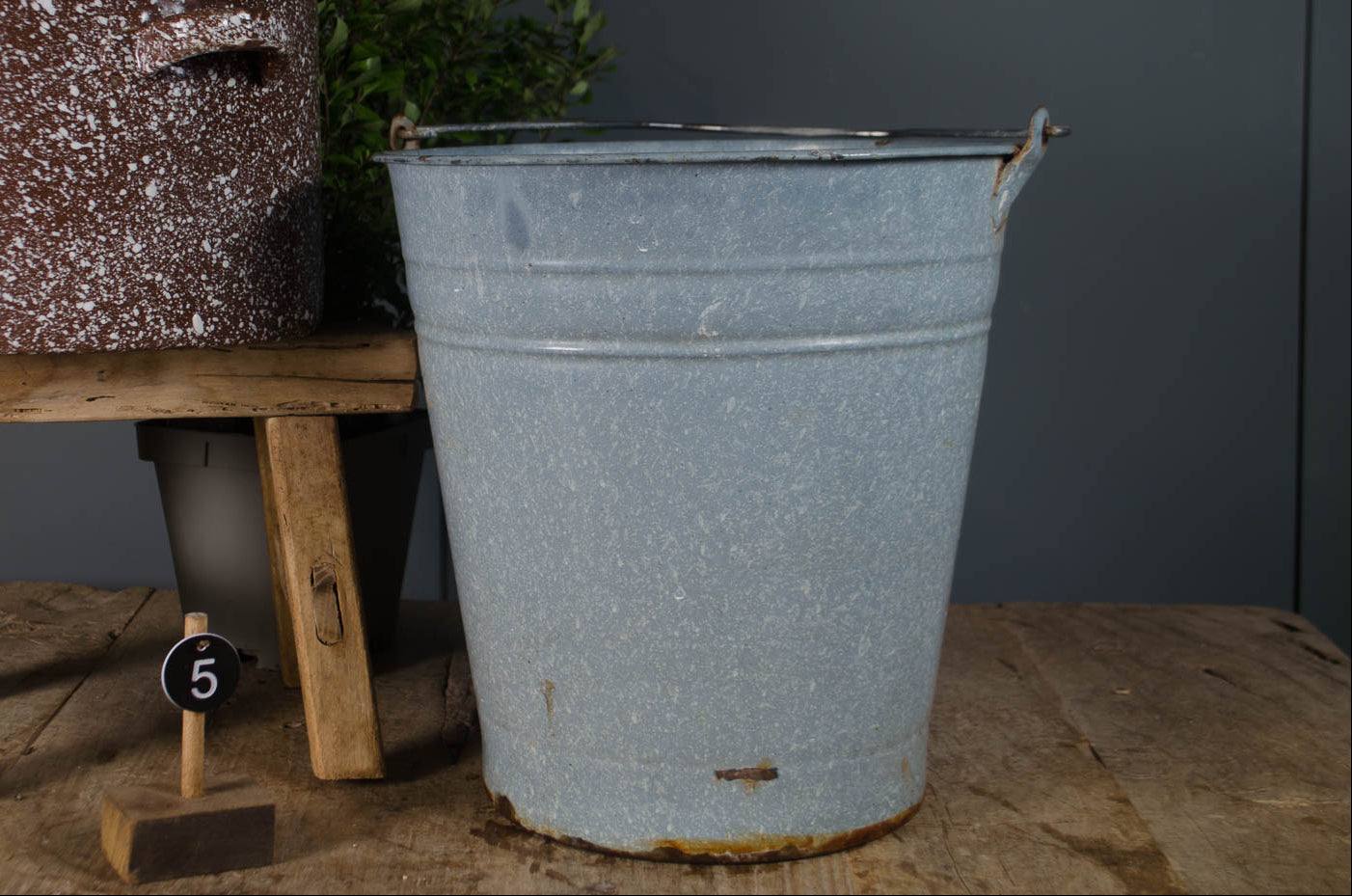 Decorative pot with flowers on a wooden stool next to a metal bucket against a dark wall.