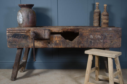 Vintage wooden table with rustic bottles against a blue wall Reclaimed wooden work bench with original vice, repurposed as a console table or desk. Ideal for hallways, studios, and industrial-style interiors.
