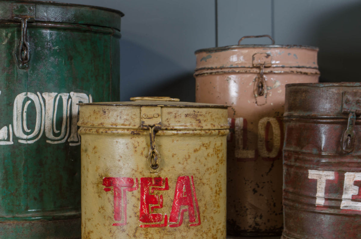 A set of vintage, distressed metal tea and flour canisters in green, cream, pink, and brown, arranged together against a dark gray background.