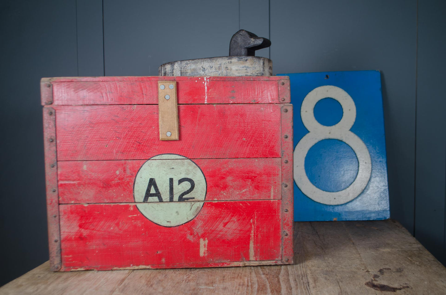 A vintage red wooden toolbox with a circular 'A12' label on the front, standing on a wooden surface against a dark gray paneled wall, with a blue sign featuring a white number 8 leaning behind it.
