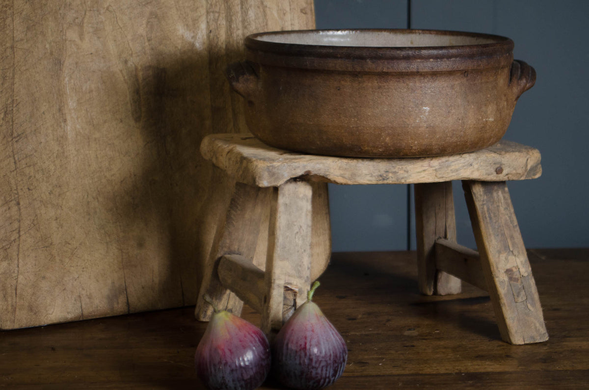 A rustic brown clay pot with side handles sits on a small wooden stool, with two purple figs on the table below and a large wooden board behind it.