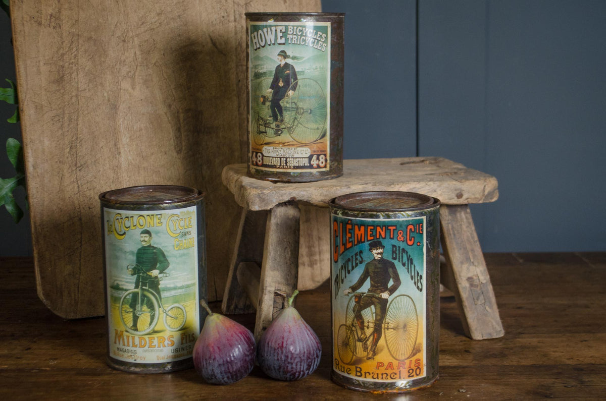 Three vintage metal tin cans with bicycle-themed labels are arranged on a rustic wooden stool and tabletop, with two purple figs in front. The background includes a wooden cutting board and a dark blue paneled wall.