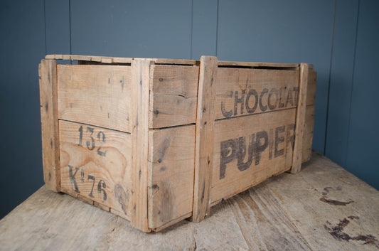 A vintage wooden crate with 'CHOCOLAT PUPIER' printed on the side, resting on a wooden surface against a dark blue paneled wall.
