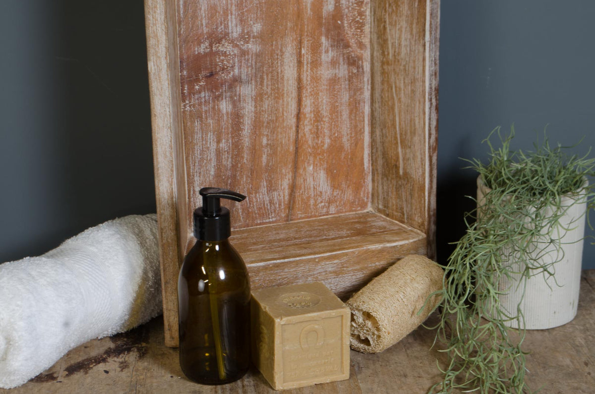 A styled product arrangement on a rustic wooden surface featuring a dark amber pump bottle, a beige rectangular bar of soap with 'LOVE' embossed, a beige textured bath sponge, a white towel, and a potted trailing green plant against a dark gray wall and distressed wood panel backdrop.