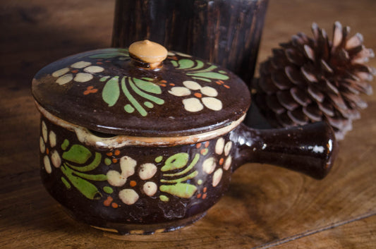 A dark brown glazed ceramic lidded pot with hand-painted green, beige, and orange floral motifs, set on a rustic wooden table next to a pinecone.