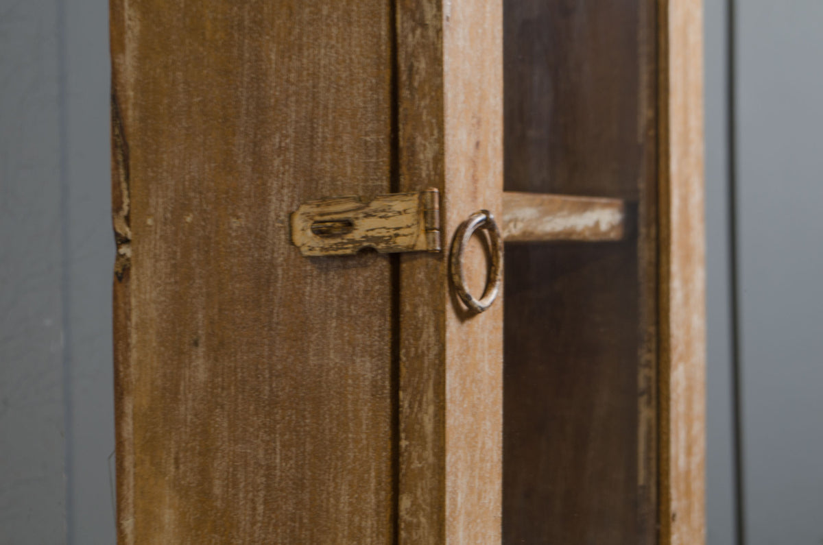 Close-up of a rustic wooden cabinet door with a distressed brass latch and ring pull on a gray background.