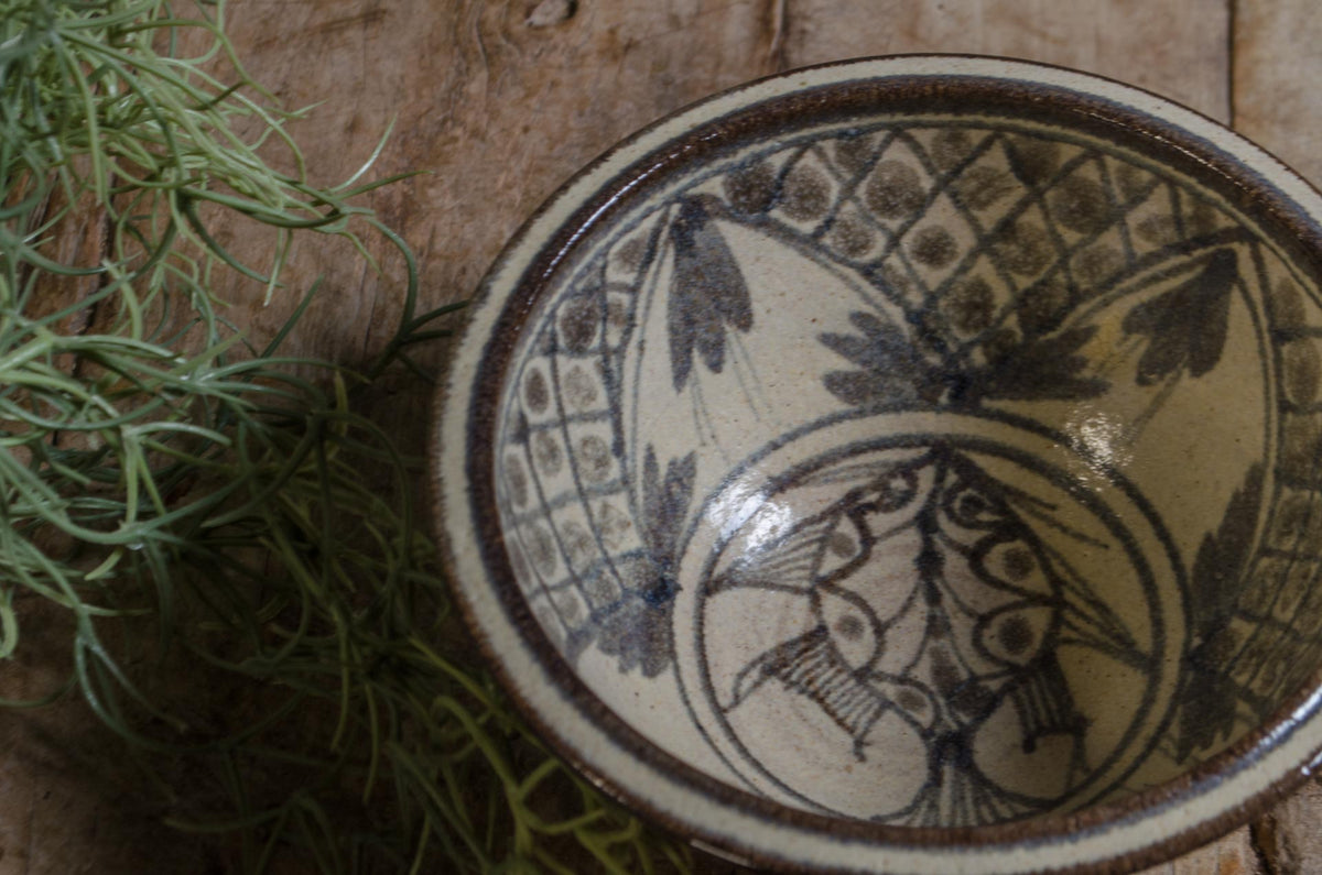 A close-up of a hand-painted ceramic bowl with a dark brown floral and fish pattern on a cream-colored glaze, styled on a rustic wooden surface next to green moss.