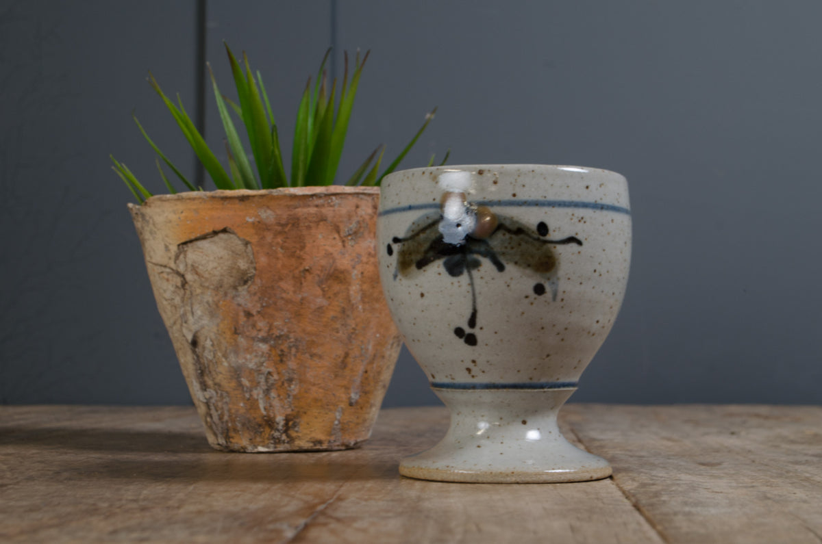 A handmade ceramic egg cup with a speckled cream glaze, blue stripe, and black painted floral motif, displayed next to a rustic terracotta planter on a wooden surface against a dark gray background.