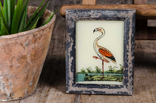 A small framed vintage-style illustration of a flamingo standing in water, displayed on a rustic wooden surface next to a potted green plant.