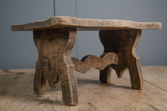 Primitive French wooden stool with weathered grey finish and carved rustic base on wooden table