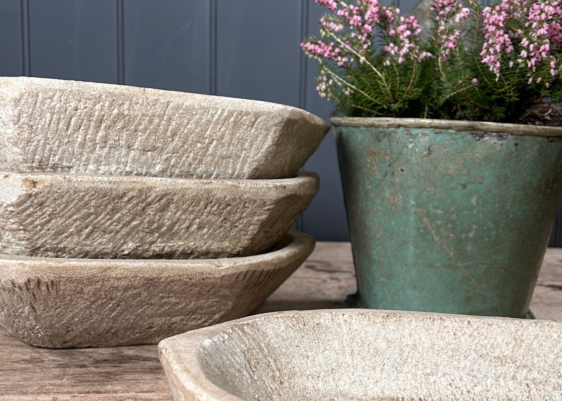 
Three stone bowls and a green pot with pink flowers on a wooden surface against a blue wall.