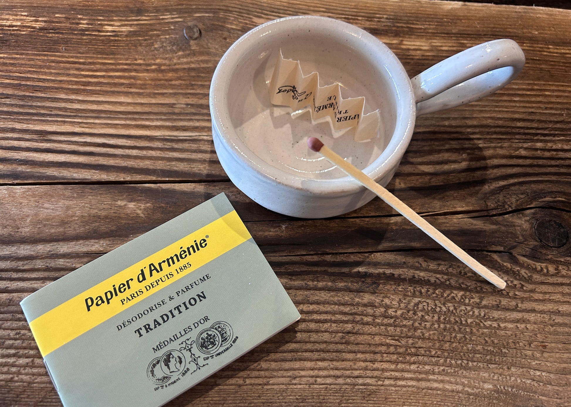 
Papier d'Armenie paper burning with a matchbox and a cup on a wooden surface.  Armenian paper insence natural fragrance for your home
