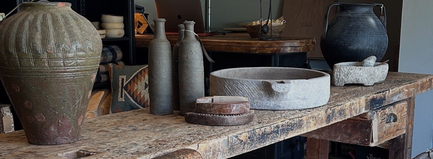 A rustic wooden workbench or table in a styled interior, with large ceramic vases, bottles, a stone bowl, and woven pendant lights around it.