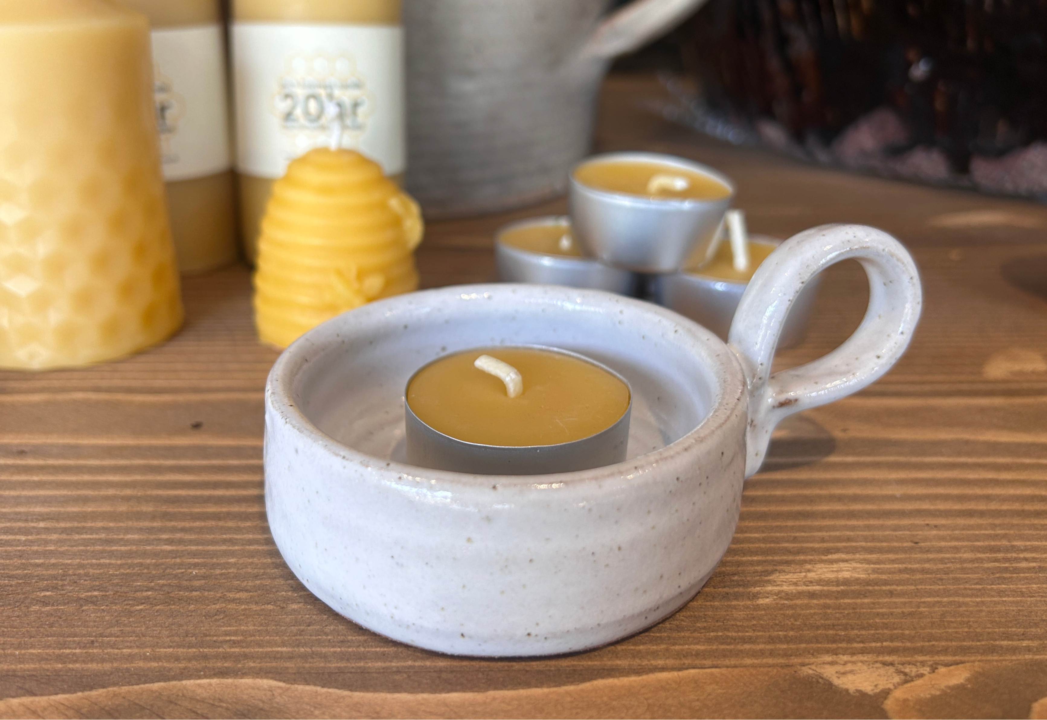 Ceramic candle holder with a lit tealight candle on a wooden surface, surrounded by other candles and containers.
