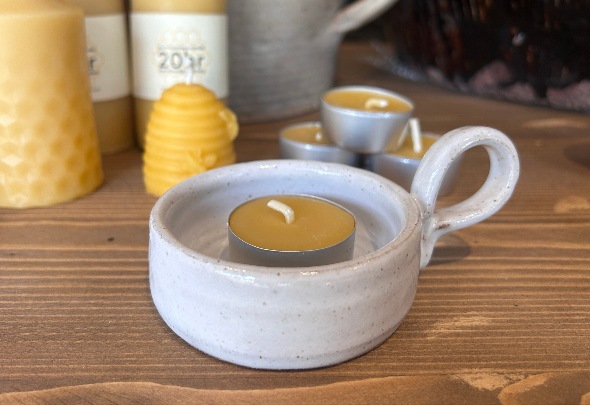 
Ceramic candle holder with a lit tealight candle on a wooden surface, surrounded by other candles and containers.