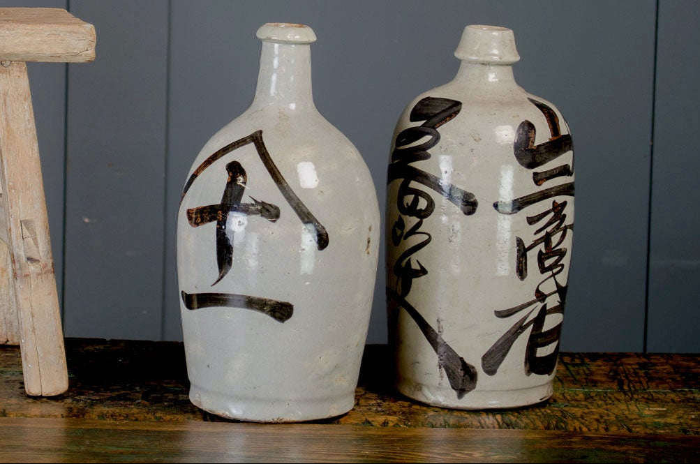 Three ceramic bottles with black characters on a wooden stool against a gray wall.