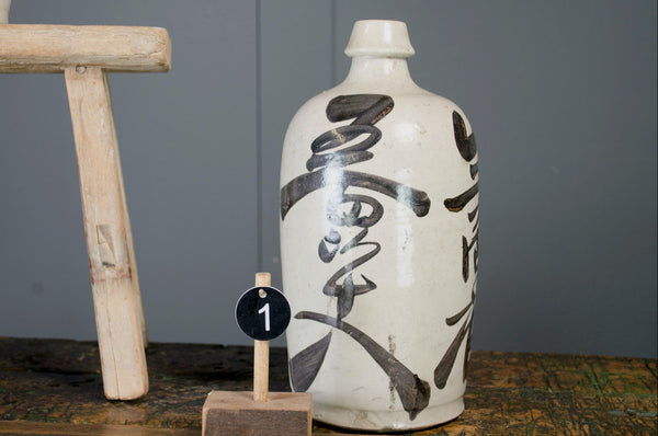 Ceramic japanese antique sake bottle with black script on a wooden stand against a grey wall.