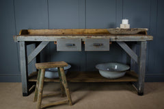 Vintage-style wooden workbench with drawers, a stool, and bowls against a dark gray wall.