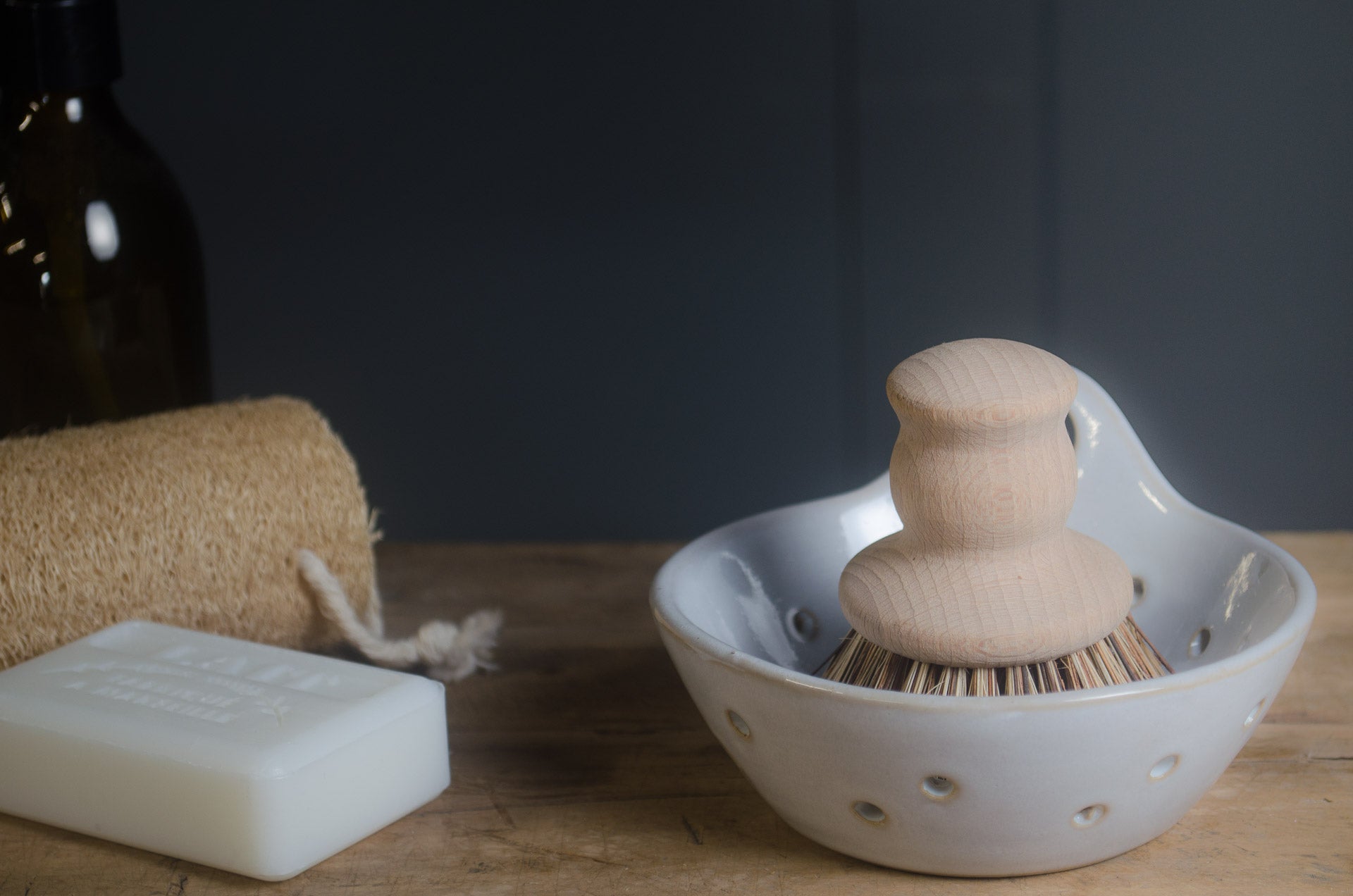 White ceramic whisk holder with wooden whisk, white soap bar, and brown towel on a wooden surface.