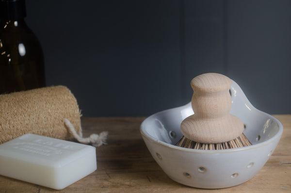 White ceramic whisk holder with wooden whisk, white soap bar, and brown towel on a wooden surface.