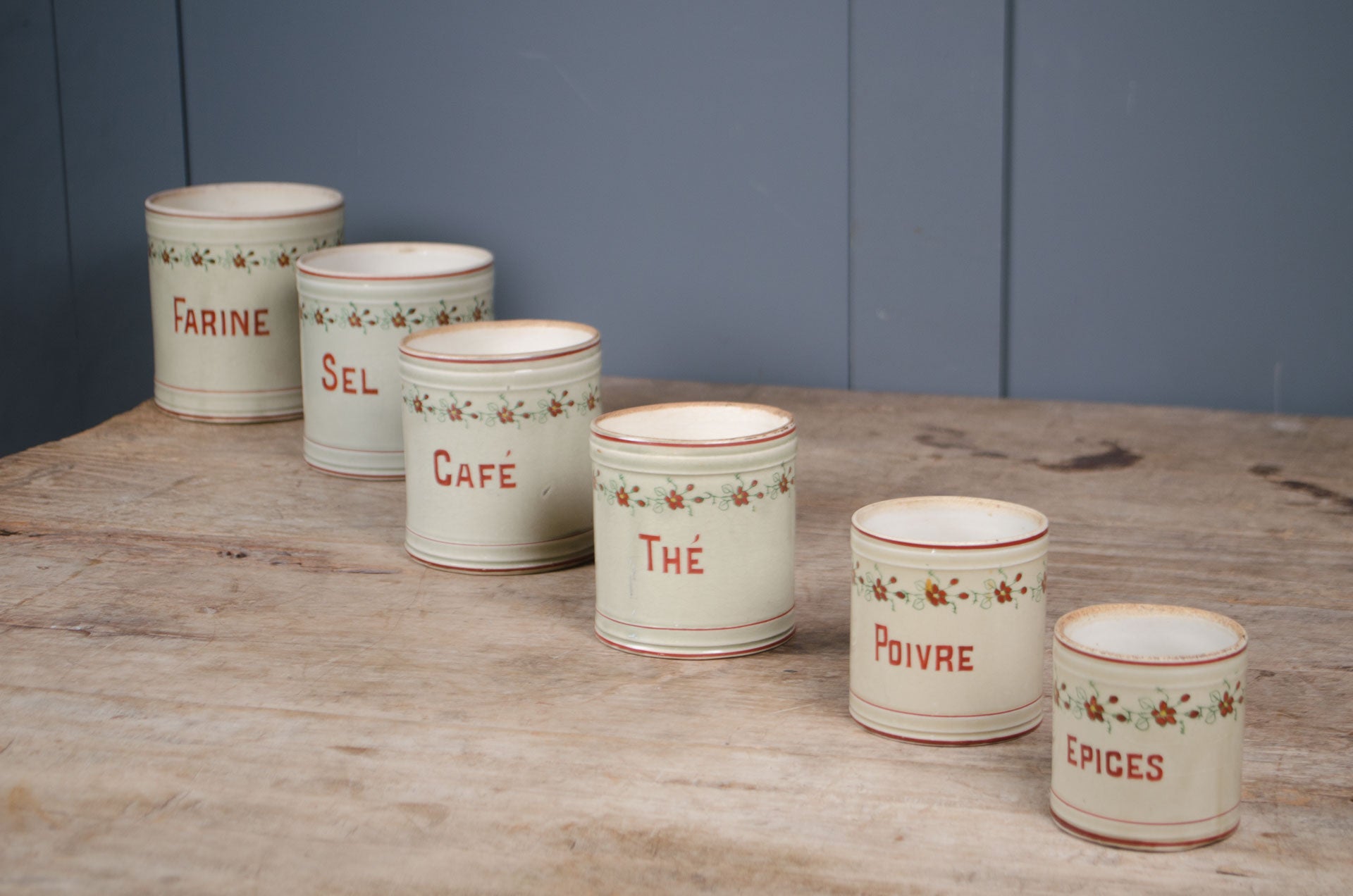 
Set of five vintage French ceramic kitchen canisters with hand-painted floral decoration and red lettering displayed on a wooden table.