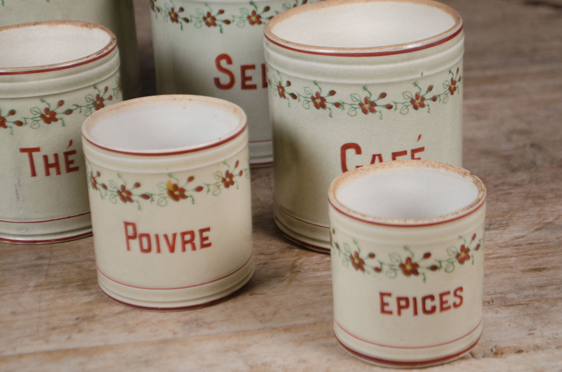 
Set of five vintage French ceramic kitchen canisters with hand-painted floral decoration and red lettering displayed on a wooden table.