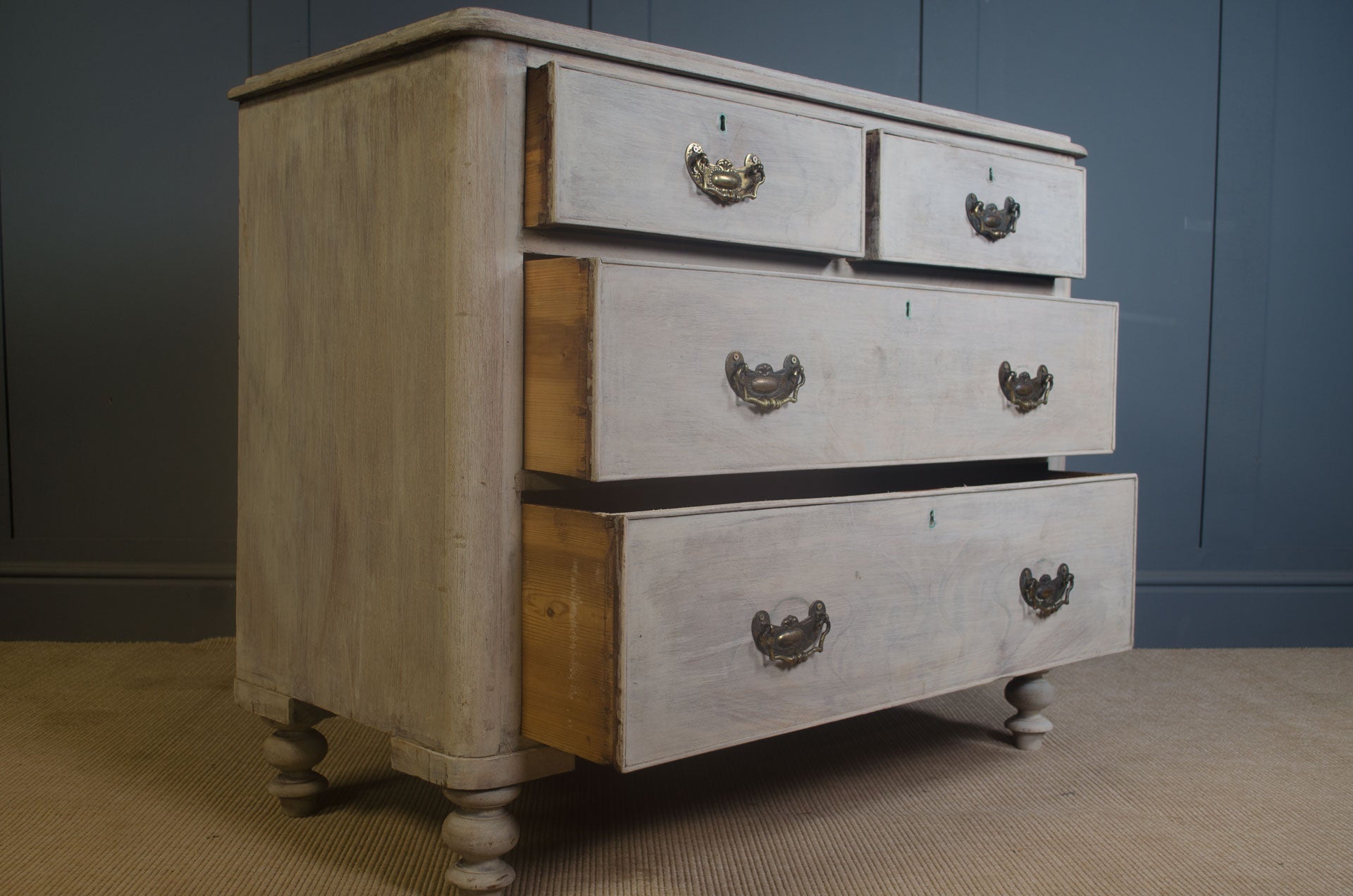 Edwardian pine chest of drawers with bleached finish, antique brass handles, and turned feet against a dark wall background