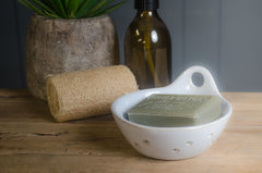 Bar of French soap in a white ceramic dish on a wooden surface with a plant and bottle in the background.