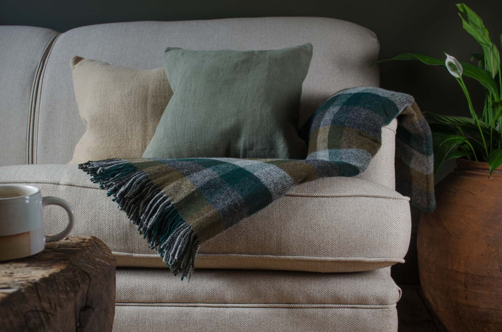 
Beige sofa with a plaid blanket, cushions, and a mug on a wooden table in a cozy room.