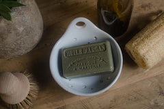 Bar of soap labeled 'Huile d'Argan' in a white dish on a wooden surface with a plant and sponge.