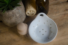 White colander with holes on a wooden surface with a plant, sponge, and bottle in the background.