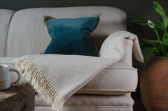 Beige sofa with a blue pillow and white blanket, next to a wooden side table with a mug and a plant and beige herringbone wool blanket