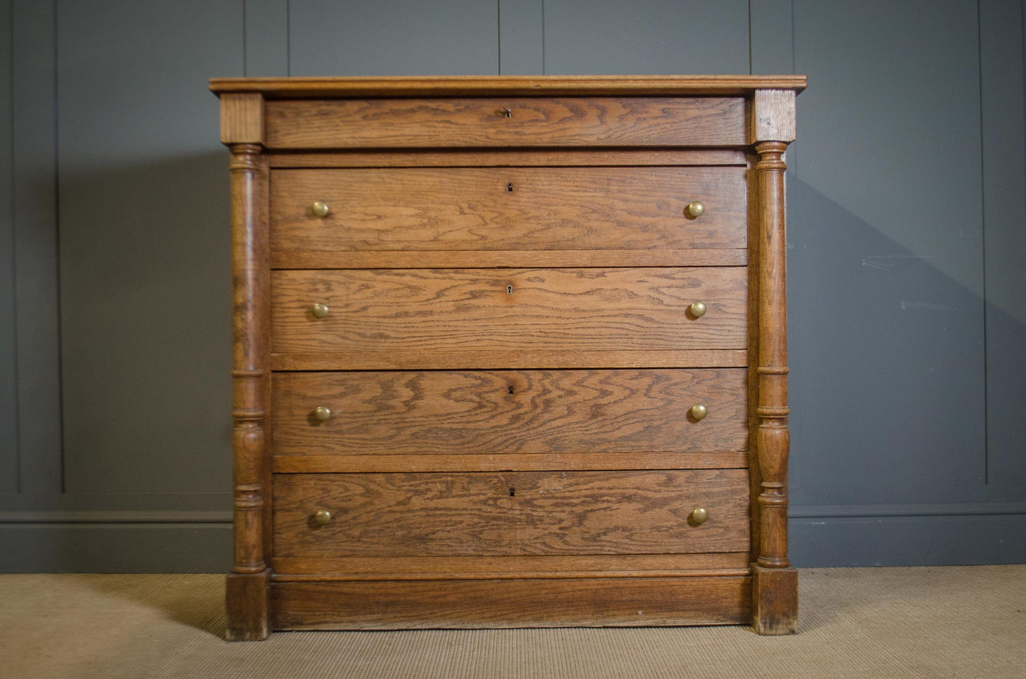 A five-drawer wooden chest of drawers with turned legs and brass knobs, set against a dark gray paneled wall and beige carpet.