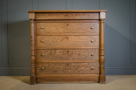 A five-drawer wooden chest of drawers with turned legs and brass knobs, set against a dark gray paneled wall and beige carpet.