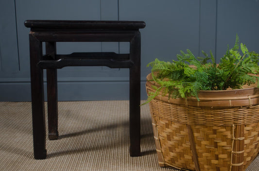 Wicker planter with plants next to a dark wooden stool against a blue wall.