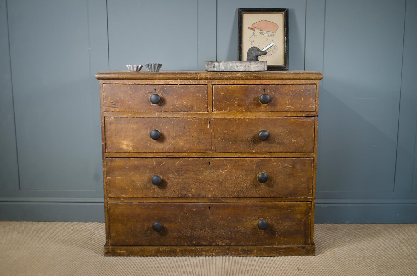 A vintage wooden chest of drawers with five drawers and dark round knobs, set against a blue paneled wall with a framed picture on top.