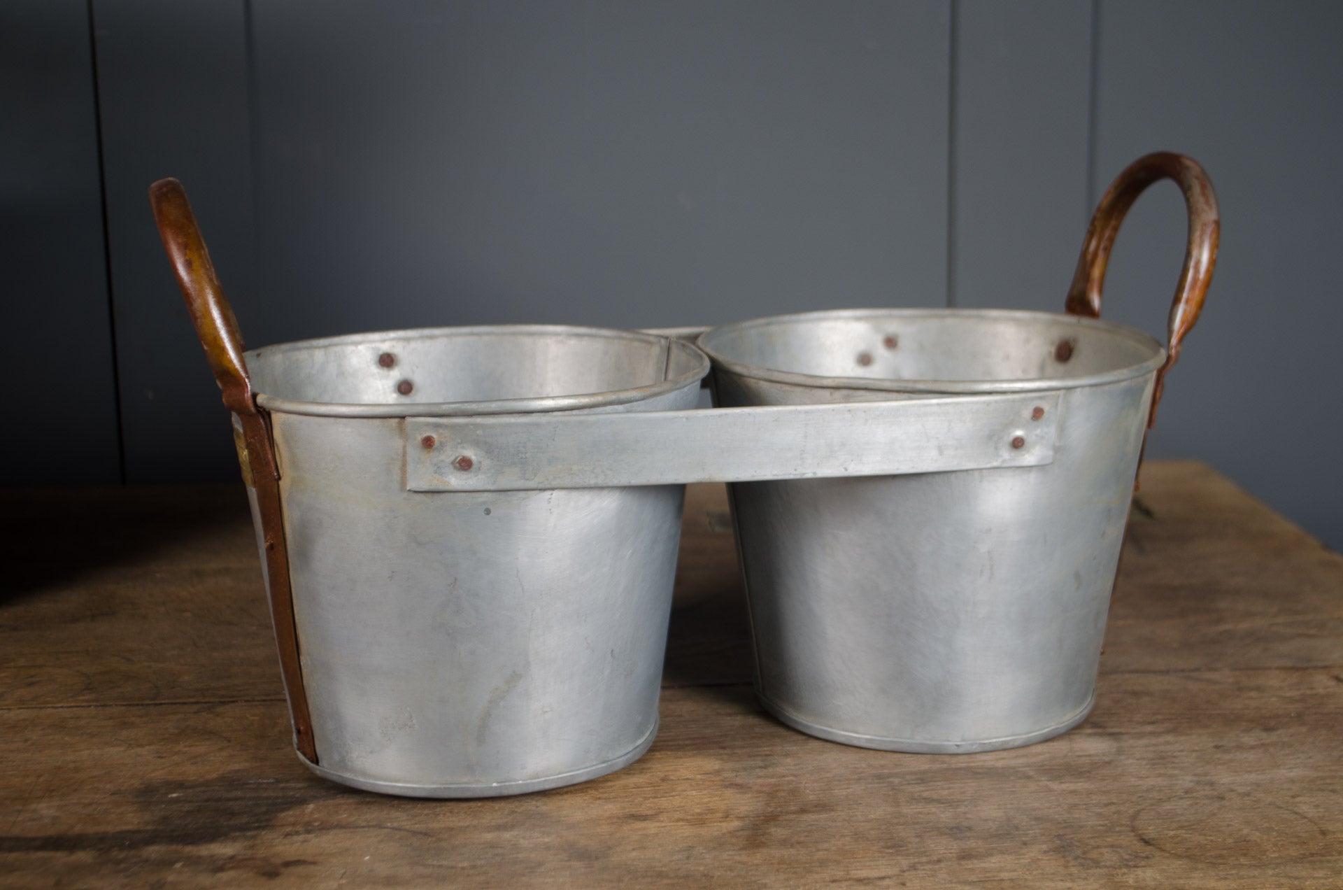 
Millwards Two metal buckets with wooden handles on a wooden surface against a gray wall.