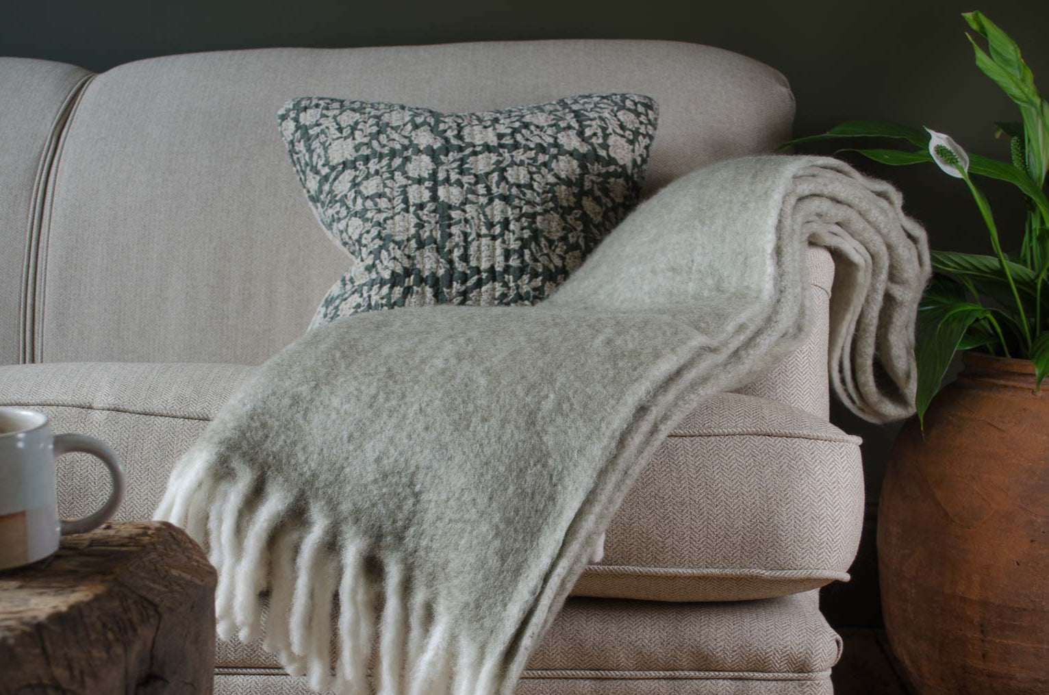 
Beige sofa with a cosy beige lambswool blanket and green cushion, wooden coffee table, and potted plant in a cozy living room setting.