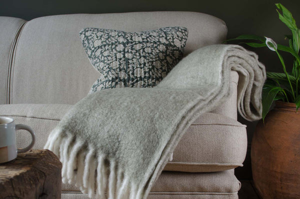 Beige sofa with a cosy beige lambswool blanket and green cushion, wooden coffee table, and potted plant in a cozy living room setting.