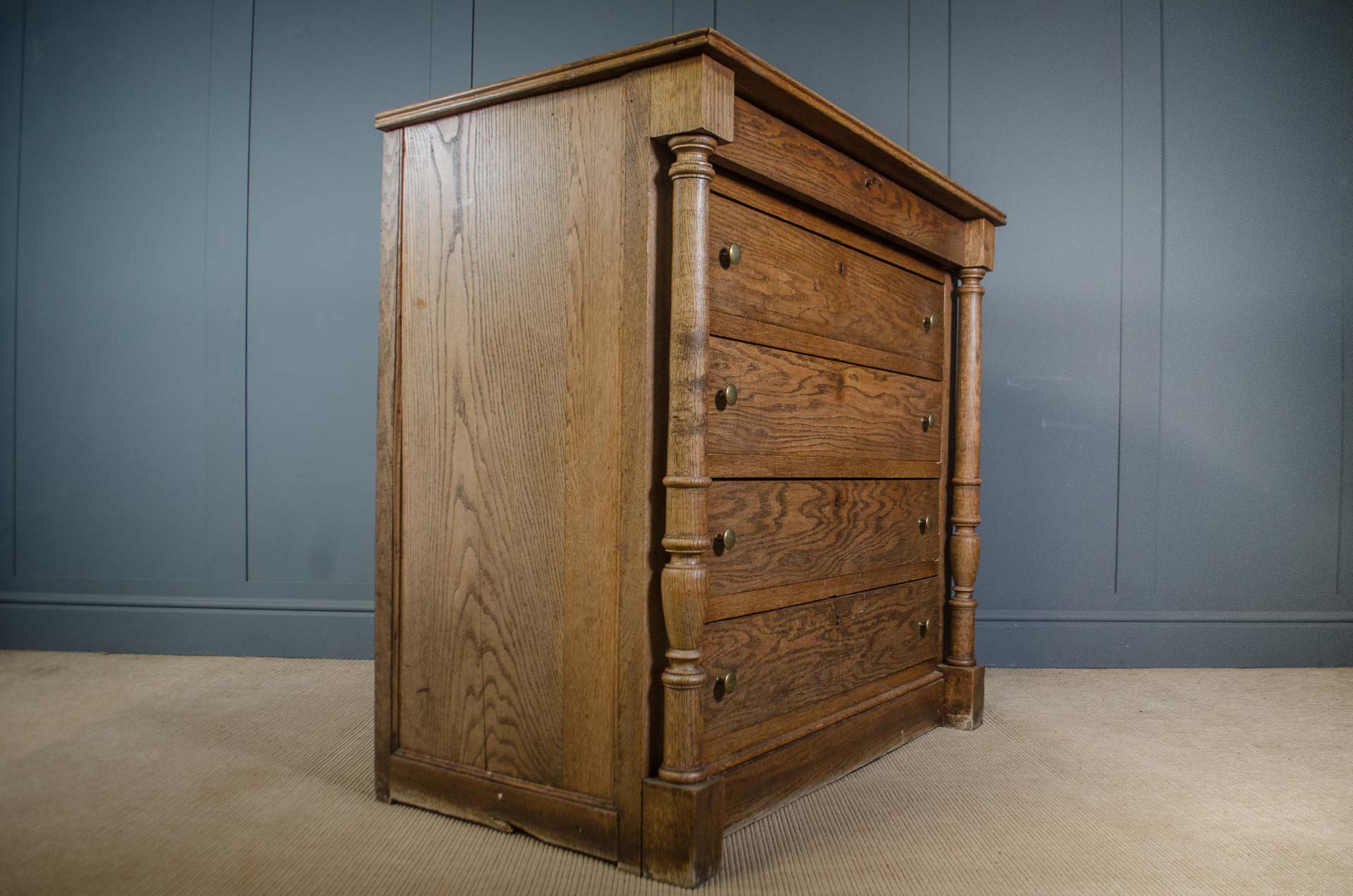 
A vintage five-drawer wooden dresser with turned legs and brass knobs, set against a blue paneled wall and beige carpet.