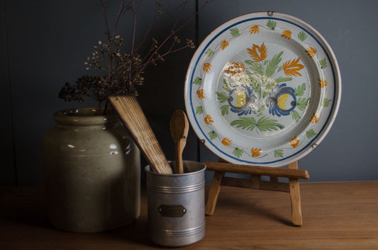 A round vintage enamel plate with a floral design, displayed on a small wooden stand on a wooden table against a dark paneled wall.