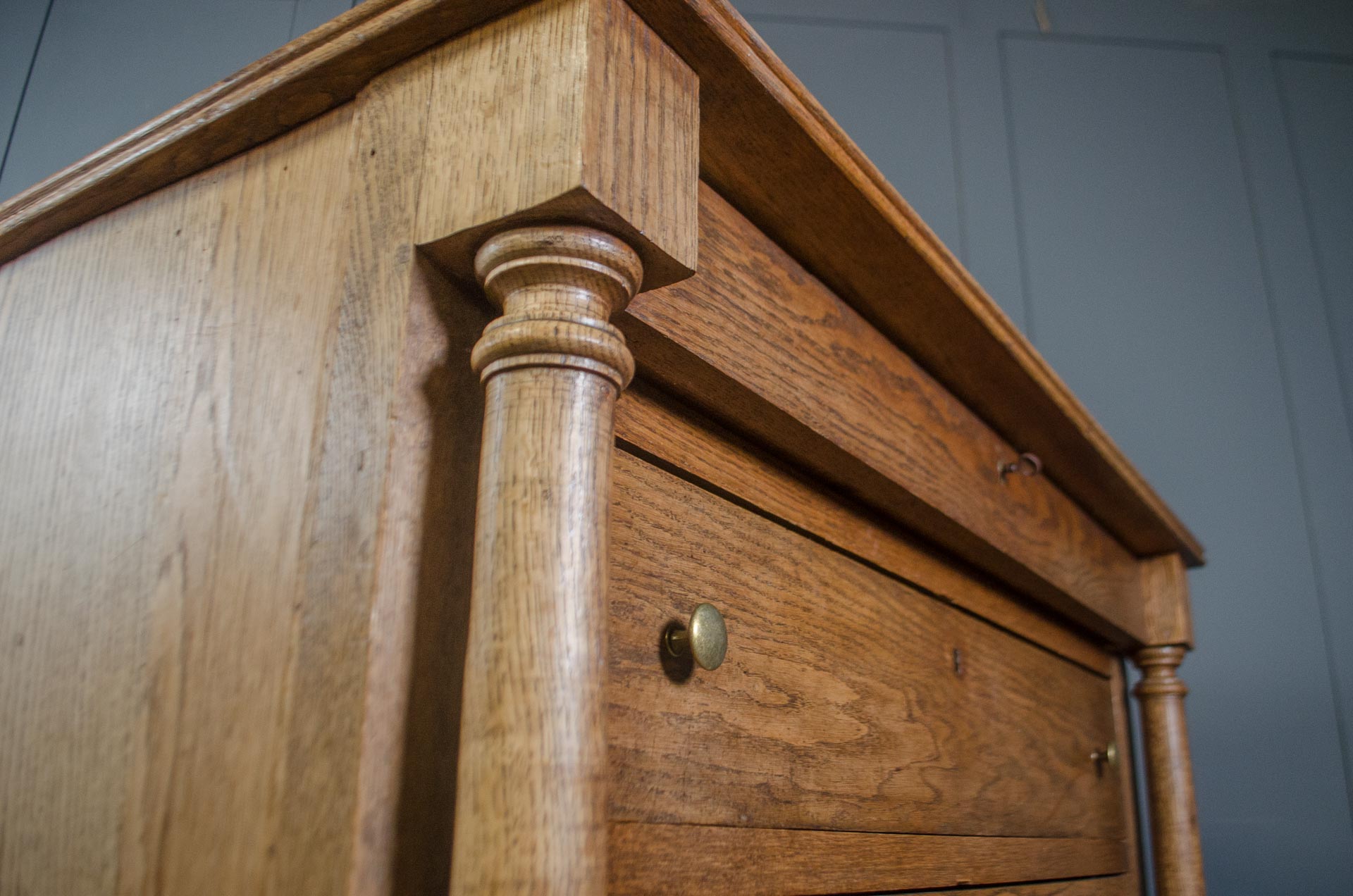
Close-up of a rustic wooden chest of drawers with a turned column leg and a brass drawer pull, set against a dark gray paneled wall.