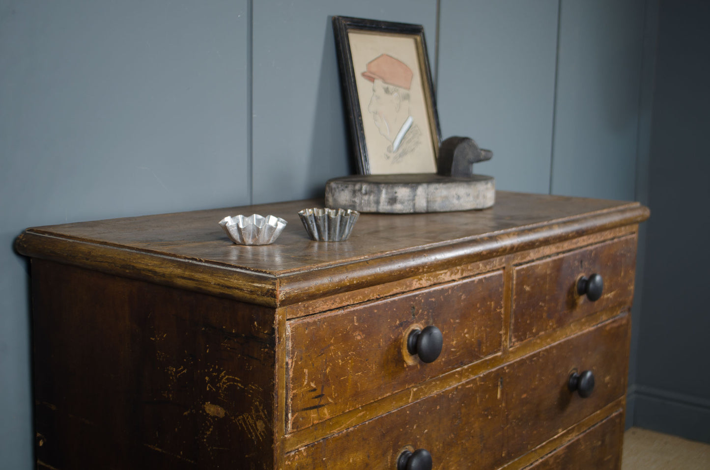 An antique wooden dresser with a distressed brown finish and black knobs, styled with small silver bowls and a framed picture against a blue paneled wall.