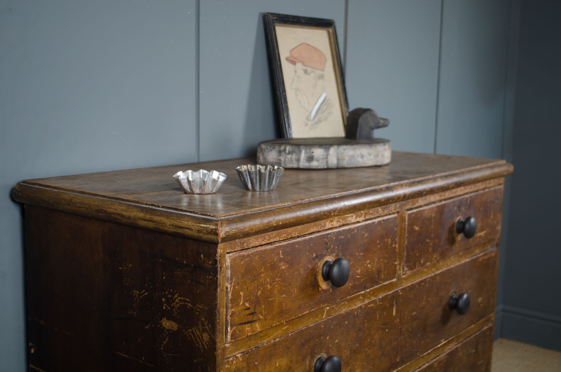 
An antique wooden dresser with a distressed brown finish and black knobs, styled with small silver bowls and a framed picture against a blue paneled wall.