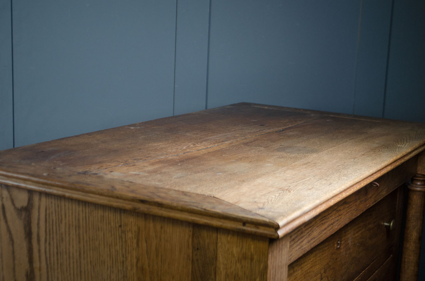 A close-up of a rustic wooden chest of drawers or side table, showing the top surface and front drawer front against a blue paneled wall.