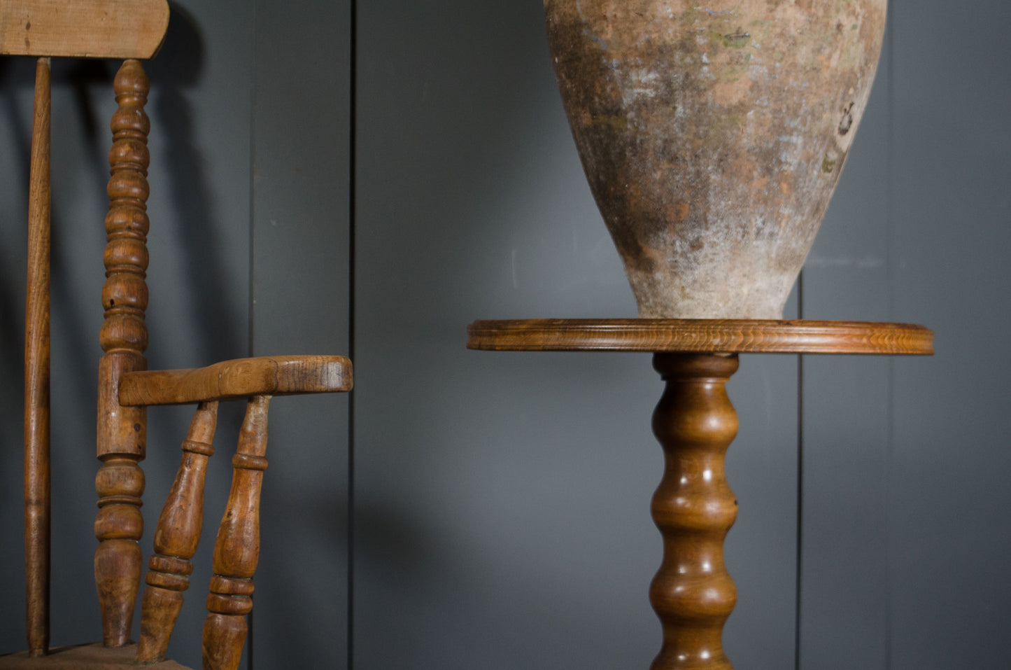 A rustic brown wooden pedestal table with a round top and turned spindle legs, holding a large textured beige vase, set against a dark gray paneled wall.